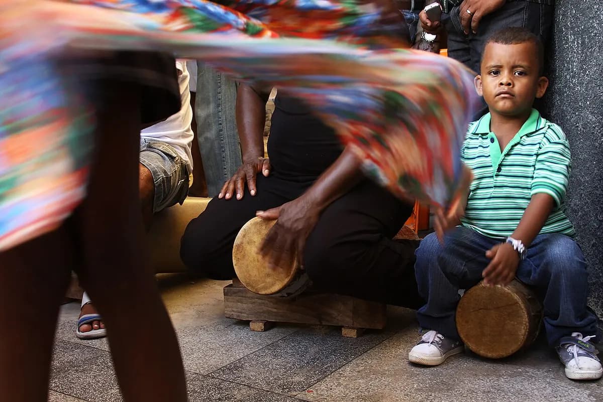 Coreiro tocando tambor de madeira apoiado no chão enquanto a saia estampada de uma coreira gira à frente, com um menino sentado ao lado