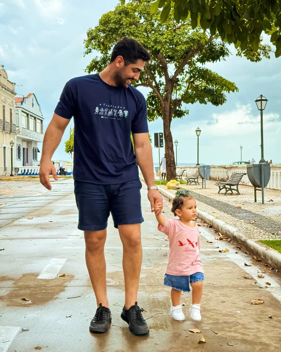 Pai e filha de mãos dadas em uma praça arborizada de São Luís, os dois com camisetas da Kambada
