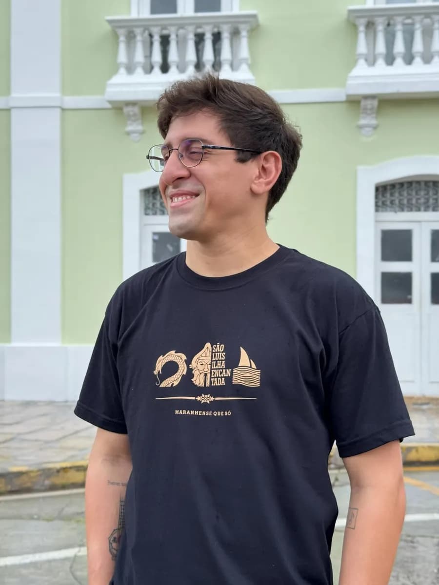 Rapaz sorrindo com a camiseta preta São Luís Ilha Encantada, em frente a um casarão verde do centro histórico